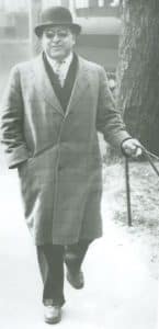 A man in an overcoat walks towards he camera. He is a white cane user.