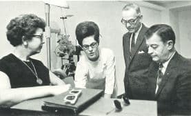 Black and white photo of two women on left and 2 men on right as they examine a pair of glasses.