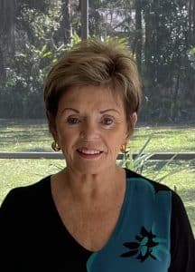 Ginny Martin - a woman standing outside smiling. She has short hair and is wearing earrings and a green and black shirt.