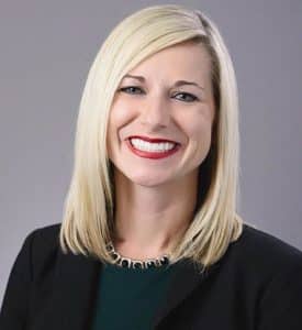 Lyndi Sheets - Smiling woman with platinum blond hair. She is wearing bright red lipstick, a dark green shirt, and black blazer. She is standing in front of a gray background.