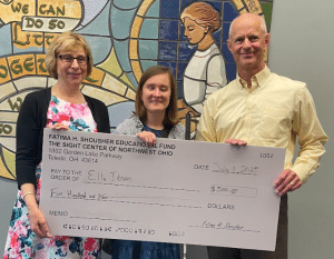 Fatima Shousher (left), Ella Ibsen (center), and Tim Tegge, Executive Director (right), stand smiling and holding an oversized ceremonial check. Fatima is wearing glasses, a black cardigan, and a pink, blue, and white floral dress. Ella has shoulder-length light brown hair and is wearing a gray cardigan over a blue dress. Tim is bald and dressed in a pale yellow button-down shirt with brown trousers. The check is from the "Fatima H. Shousher Educational Fund" at The Sight Center of Northwest Ohio, made out to Ella Ibsen for $500, dated July 1, 2025. Behind them is a colorful stained glass mural with stylized human figures and text reading “WE CAN DO SO MUCH TOGETHER.”