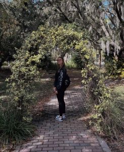 Smiling woman wearing all black and glasses standing on a brick pathway surrounded by trees and greenery.